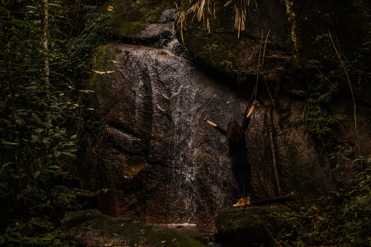 Uma mulher levanta os braços em celebração em frente a uma cachoeira caudalosa na floresta exuberante de Moeda, Brasil.