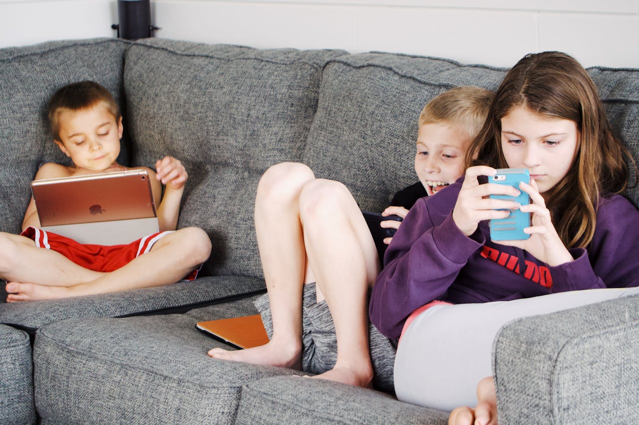 Crianças descalças positivas com roupas casuais descansando juntas em um sofá aconchegante e navegando em tablets e smartphones.