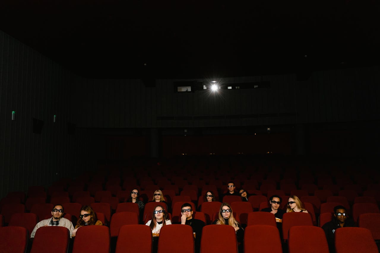 Um grupo de pessoas em um cinema assiste a um filme 3D usando óculos, sentadas em uma sala escura.